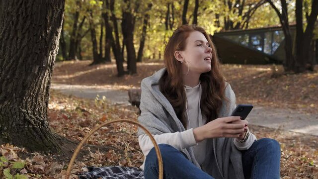 Beautiful young woman sits on blanket in the park and uses phone at fall on a sunny day