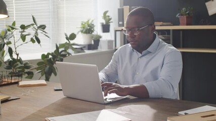 Arc shot of professional African American businessman typing on laptop during workday while sitting at desk in office - Powered by Adobe