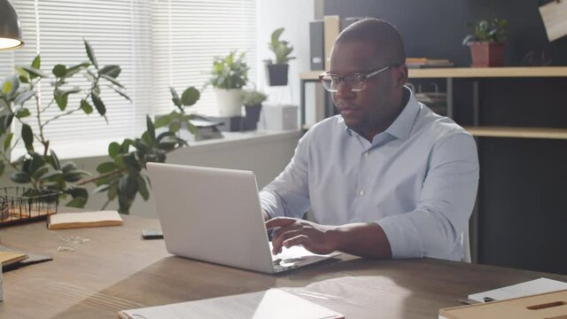 Arc shot of professional African American businessman typing on laptop during workday while sitting at desk in office