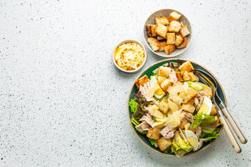 Fresh Caesar salad with lettuce salad, chicken breast, boiled eggs and croutons in ceramic bowl with dressing on the side on white table. Classic healthy salad, top view, space for text