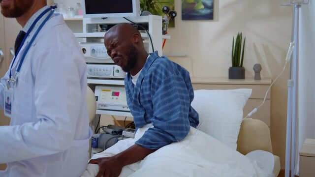 Indifferent doctor sitting on man's leg lyinghospital bed mistreating patient. Afro-american sick guy crying with pain. Incompetence. Comical scene. Problem concept.