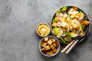 Fresh Caesar salad with lettuce salad, chicken breast, boiled eggs and croutons in ceramic bowl with dressing on the side on gray table. Classic healthy salad, top view, space for text