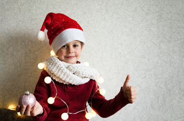 Festive little boy in Santa hat smiling at the camera holding a New Year's ball. Child on a light background with free space for your inscription.