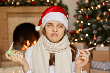 Unhappy and unhealthy woman posing in decorated living room wit thermometer and inhaler in hands, being disappointed to be ill during Christmas holidays, wearing santa hat and scarf.