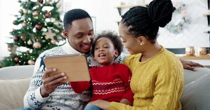 Portrait of happy African American little kid sitting on sofa in decorated room near new year tree with mom and dad and tapping on tablet while playing. Family holidays. Merry Christmas concept