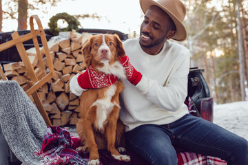 African American man sits with a dog in the trunk of a car in the winter forest. A man in a white sweatshirt.
