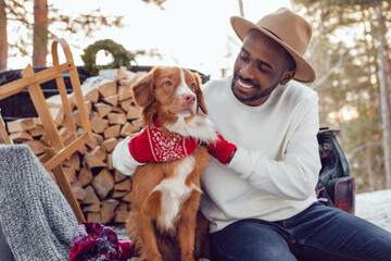 African American man sits with a dog in the trunk of a car in the winter forest. A man in a white sweatshirt.