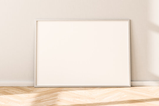 Horizontal Empty Picture Frame On Parquet Floor Leaning Against Bright Wall. Sunlight Flooding In From The Left.