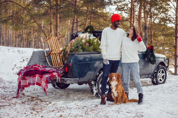 Multiracial couple in white sweatshirts stand with a dog near a car in the forest.