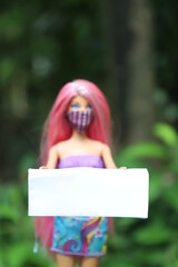 A pink haired doll wearing a mask and holding a white clean banner to add or write text in nature