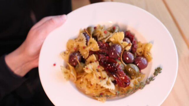 woman hands putting a white plate of farfalle pasta with sun-dried tomatoes and capers on the wooden desk 4K slowmo camera motion