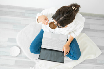 Woman in white shirt sits on floor home with laptop and drinks coffee.