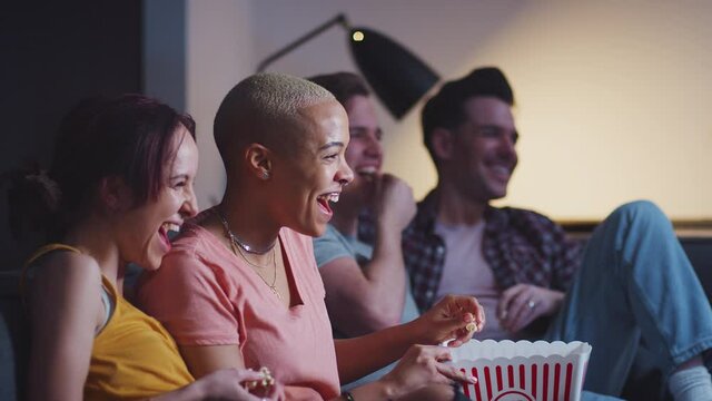 Group of gay friends sitting on sofa at home eating popcorn and laughing watching evening TV together - shot in slow motion