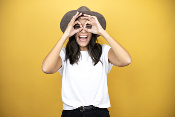 Beautiful woman wearing casual white t-shirt and a hat standing over yellow background doing ok...