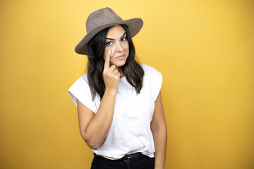 Beautiful woman wearing casual white t-shirt and a hat standing over yellow background Pointing to the eye watching you gesture, suspicious expression