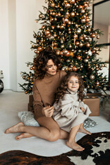 Charming mother and daughter with curly hairstyle have fun, hug and kiss at home near the Christmas tree in a white interior