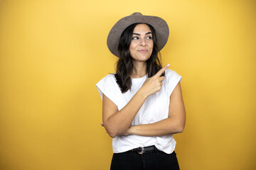 Beautiful woman wearing casual white t-shirt and a hat standing over yellow smiling happy pointing with hand and finger to the side background
