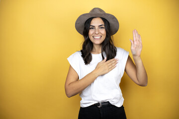 Beautiful woman wearing casual white t-shirt and a hat standing over yellow background smiling swearing with hand on chest and fingers up, making a loyalty promise oath
