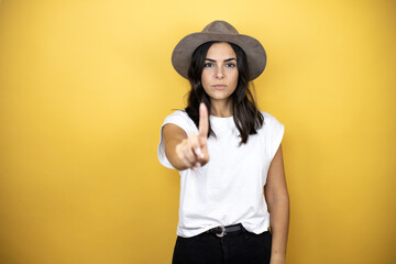 Beautiful woman wearing casual white t-shirt and a hat standing over yellow background showing and pointing up with fingers number one while is serious