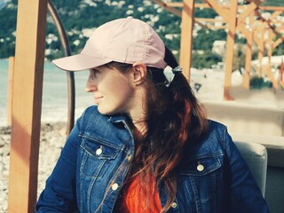 A girl with red hair in a denim jacket and a pink cap sits in a coastal cafe and looks towards the sea