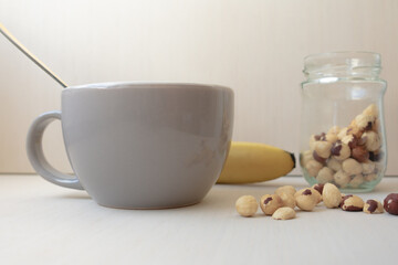 Bowl with a spoon in it, with a banana in the background, and a jar of hazelnuts and some hazelnuts on the table.