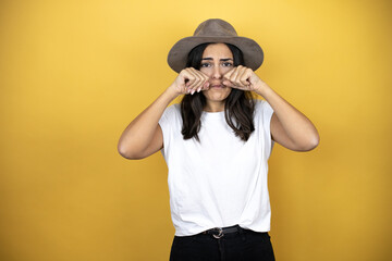Beautiful woman wearing casual white t-shirt and a hat standing over yellow background depressed and worry for distress, crying angry and afraid. Sad expression