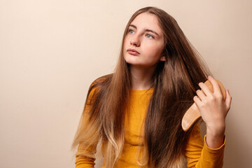 Cute teenager girl brushing her hair with wooden hair brush. The model dressed in yellow shirt and got long thick hair. Neutral color background. Beauty and haircare concept