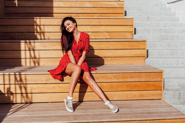 Beautiful fashionable brunette woman in summer red dress sits on the steps