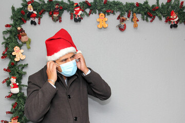 latin man with protection mask, coat and santa claus hat in christmas decoration, new normal...