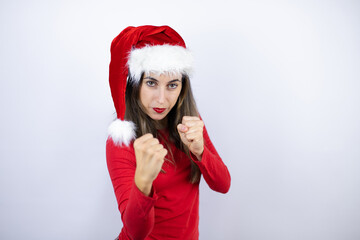 Young beautiful woman wearing a Santa hat over white background Punching fist to fight, aggressive and angry attack, threat and violence