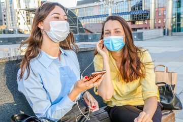 two young women in protective masks listen to music through the same headphones