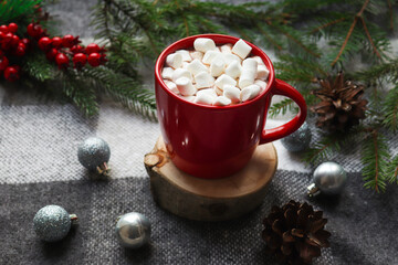 Obraz na płótnie Canvas Winter cozy home concept. Hot cocoa in a red mug with marshmallows on a wooden stand and background of gray cozy plaid, Christmas toys and pine cones. Close-up, selective focus, copy space
