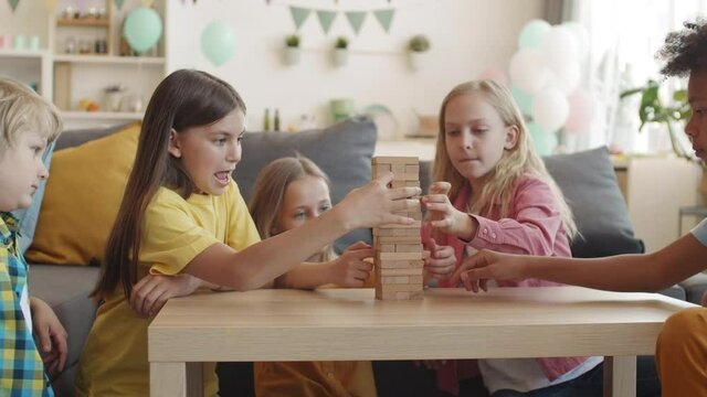 School children sitting around coffee table in living room taking out wooden bricks out of mini tower and putting them back on top. Girls and boys communicating, smiling, playing game on party