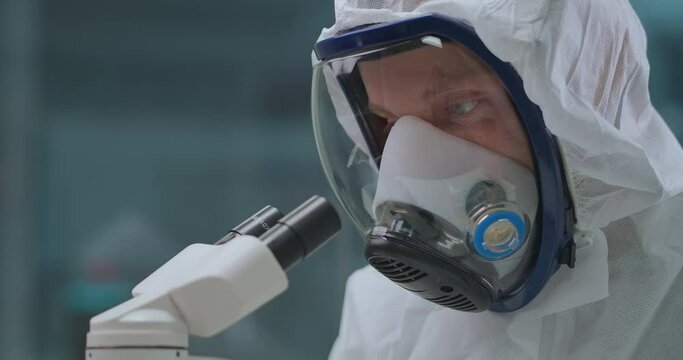 microbiologist is exploring dangerous bacteria by microscope in laboratory, protecting himself by overall and mask with respirator