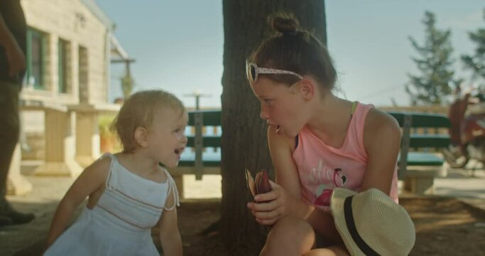 Cute little girl chatting with her older teenage sister sitting by a tree in an old european town in summer