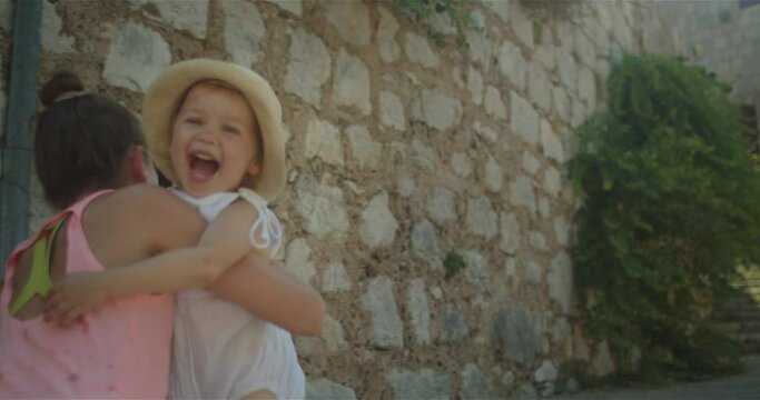 2 years old cute little girl joyfully running to her older sister in the street of an old european town, in summer