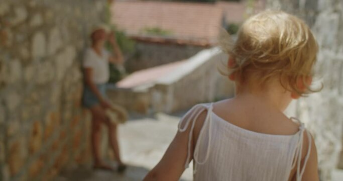Mother leaning on a stone wall waiting for her two years old cute little girl to climb down the stone steps in an old european city in summer on a sunny day