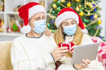 A young married couple with medical masks are sitting on the sofa in Santa hats against the background of a Christmas tree and congratulating relatives with Christmas (New Year) online using a tablet.
