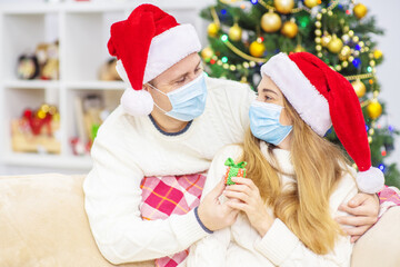 Young married couple with medical masks are sitting on the sofa wearing Santa hats and congratulating each other on Christmas by giving gifts. Holidays during the coronavirus pandemic
