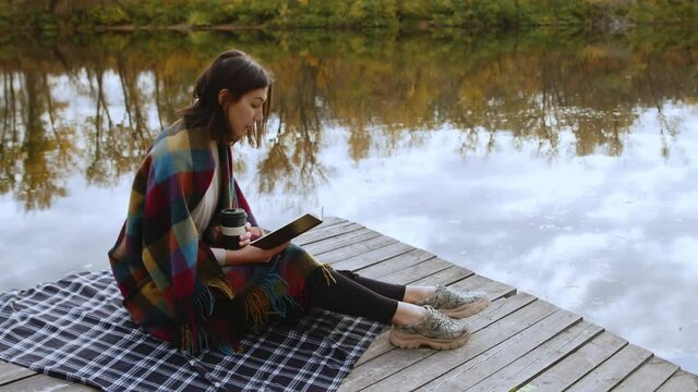 Young beautiful woman wint blanket on shoulders reads book and drinks coffee while stits po piea by the river