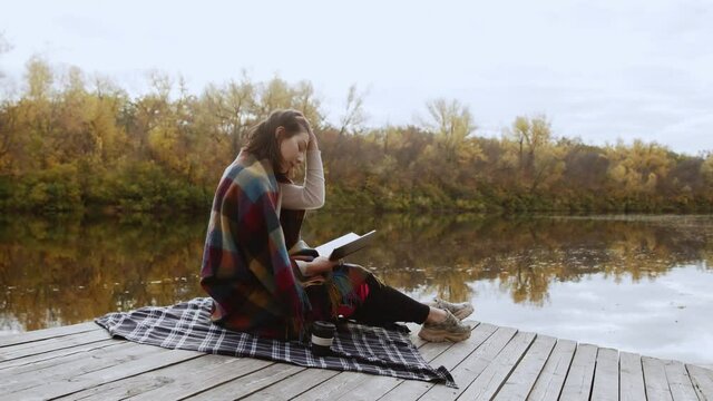 Young beautiful woman wint blanket on shoulders reads book and drinks coffee while stits po piea by the river