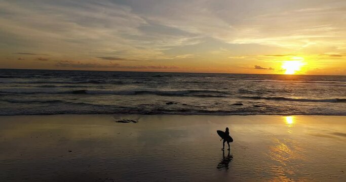 Aerial shot of surfer walking down the beach with surfboard at sunset.