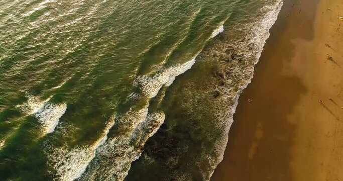 Top down drone video of waves breaking on sandy beach at sunset.
