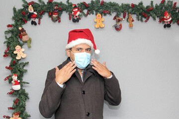 latin man with protection mask, coat and santa claus hat in christmas decoration, new normal...