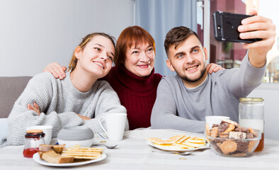 Obraz na płótnie Canvas Happy adult children taking selfie with elderly mother over cup of tea at home
