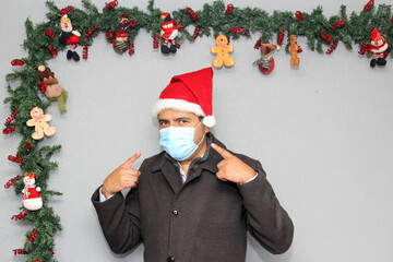 latin man with protection mask, coat and santa claus hat in christmas decoration, new normal...