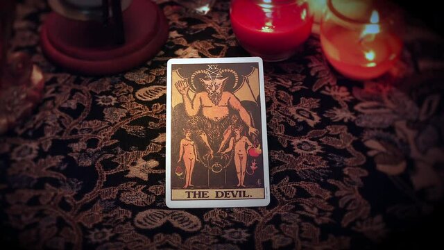 A mystical fortune teller selects a Tarot card and places it on the enchanted table. The Devil card is drawn and displayed in front of the candles and voodoo ornaments.