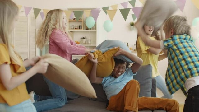 Five multiethnic school children fighting with pillows on and around couch in living room. Girls and boys, laughing, running around, having fun indoors on party