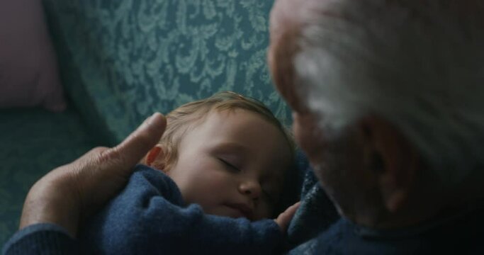 Cinematic close up shot of senior gray hair grandfather cuddling grandson baby is sleeping peacefully on arms while sitting on sofa at home. Concept:life, grandparents,love,care, generation, childhood