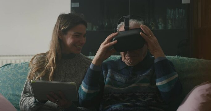 Cinematic shot of senior grandfather with vr glasses and granddaughter with tablet are having fun to use augmented reality technology together for family entertainment while sitting on sofa at home.